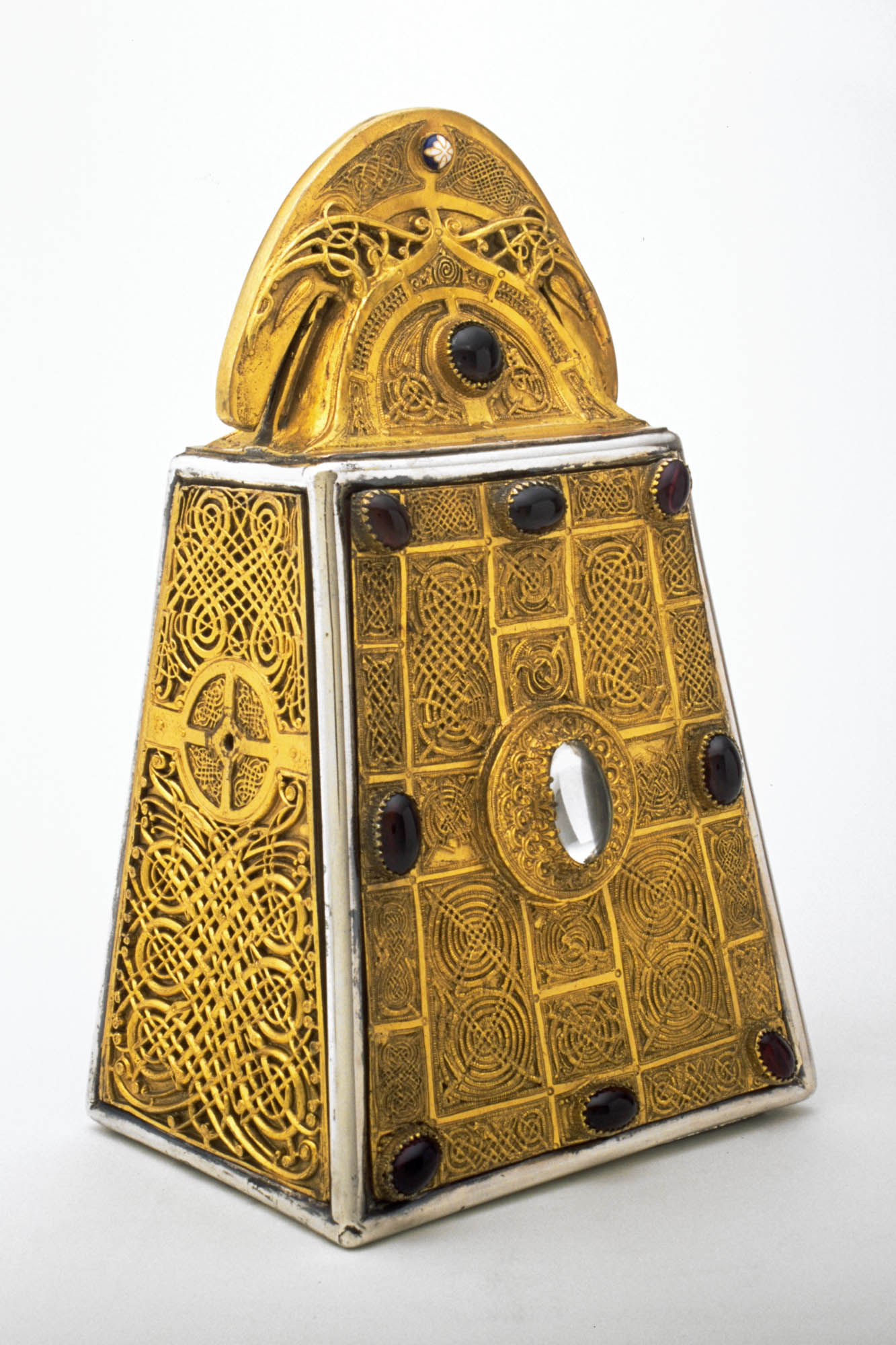 Ornate gold box in the shape of an upright trapezoidal prism covered in interlaced panels and studded with gems with semi-circular top