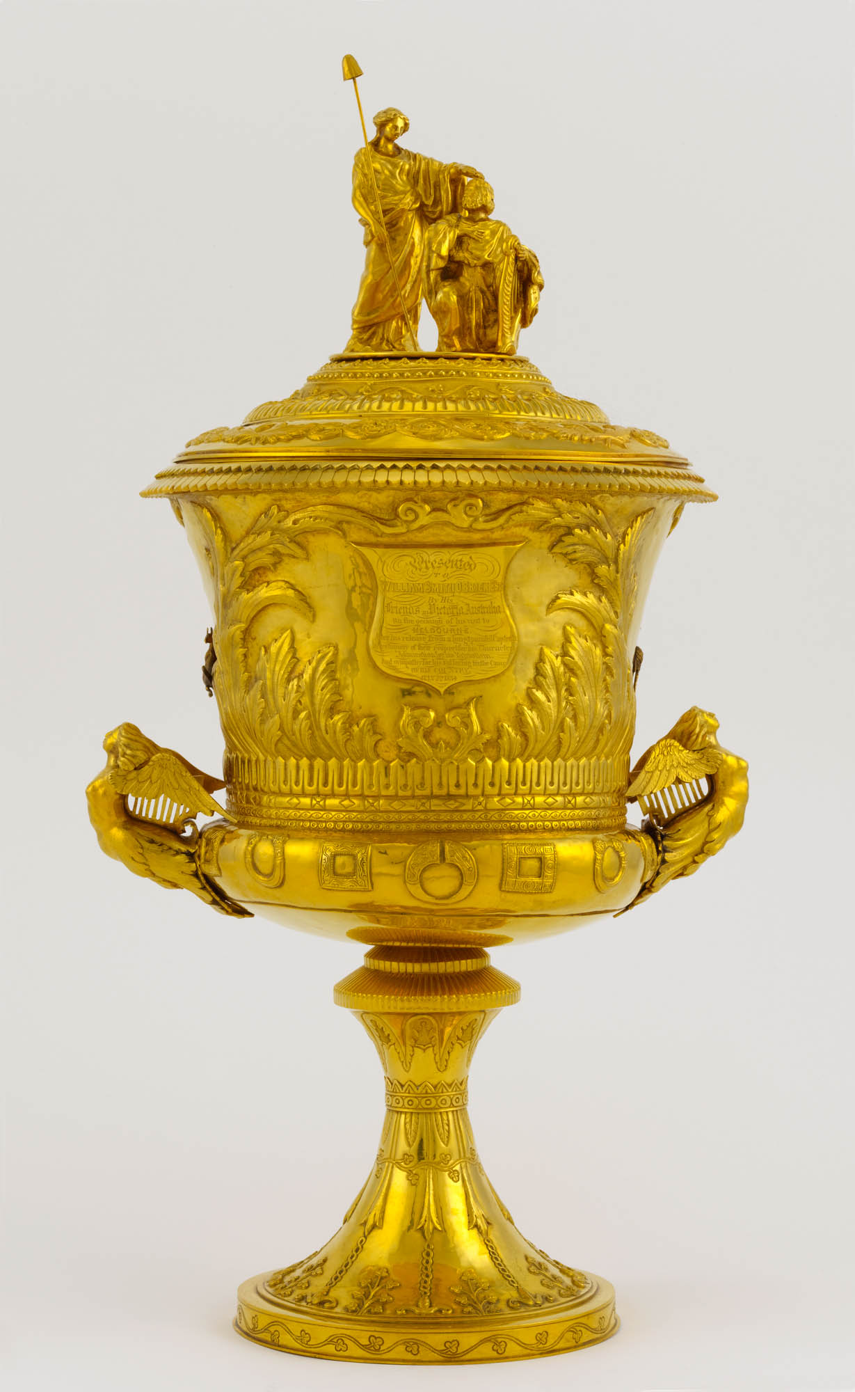 Gold cup in the shape of a trophy with two handles in the shape of winged female figures. On the lid, there are two figures of a woman touching the top of a kneeling man's head.